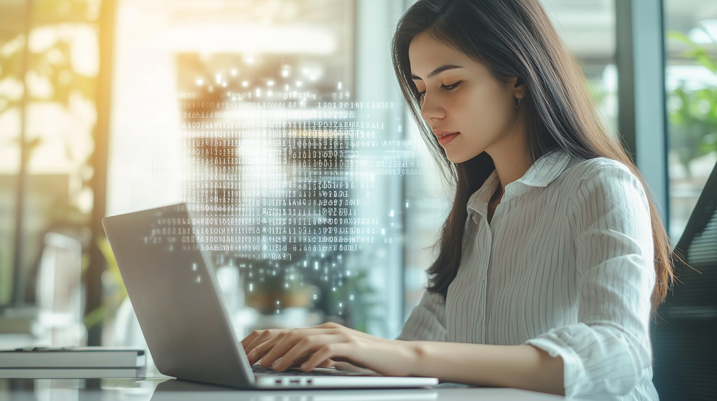 Young professional woman typing on laptop in modern office with digital code overlay, illustrating cybersecurity and real-time tech monitoring.